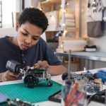 A serious teen boy uses a soldering gun to connect wires as he builds a robot at home.